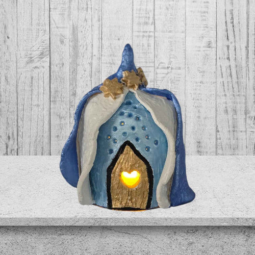 A whimsical fairy house with a blue roof sits on a sandy beach, surrounded by gentle waves and seashells.