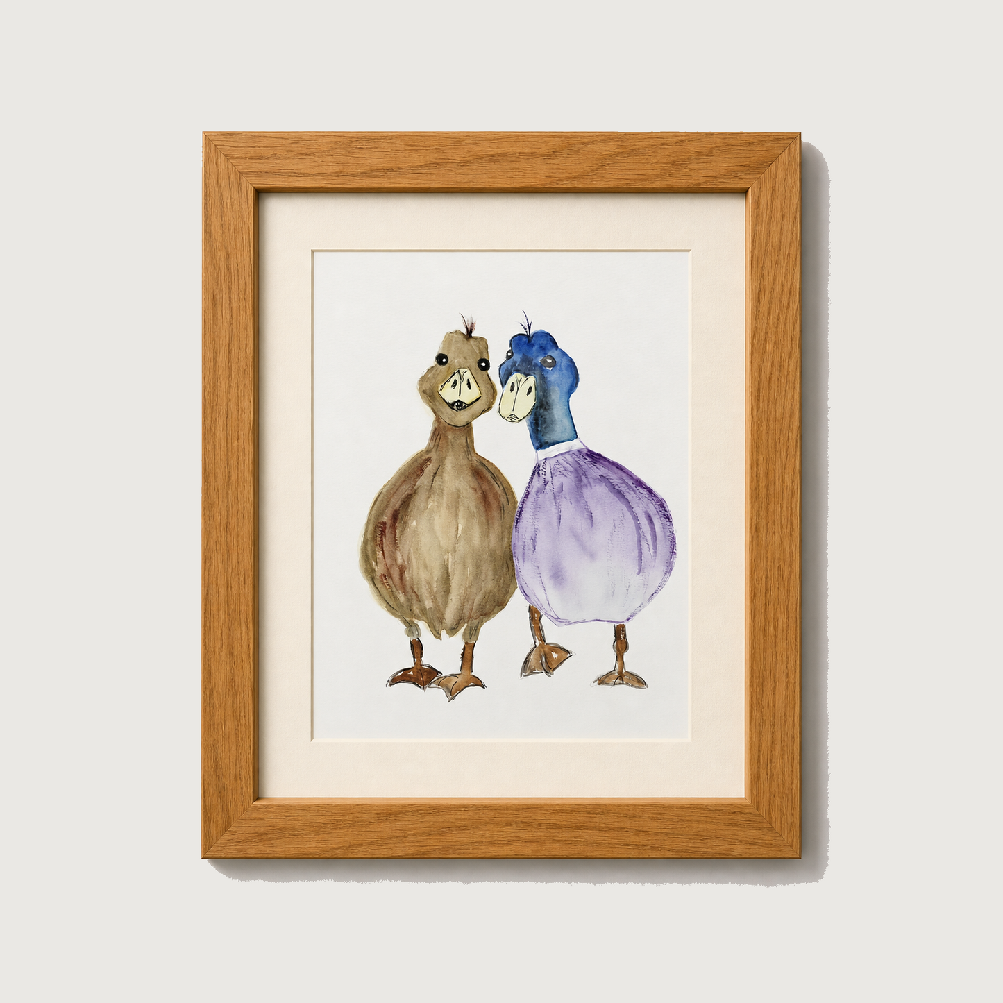 Framed watercolor art print featuring two cartoonish ducks standing side by side, one brown and one blue and purple, set in a light wood frame with a white mat against a light gray wall.