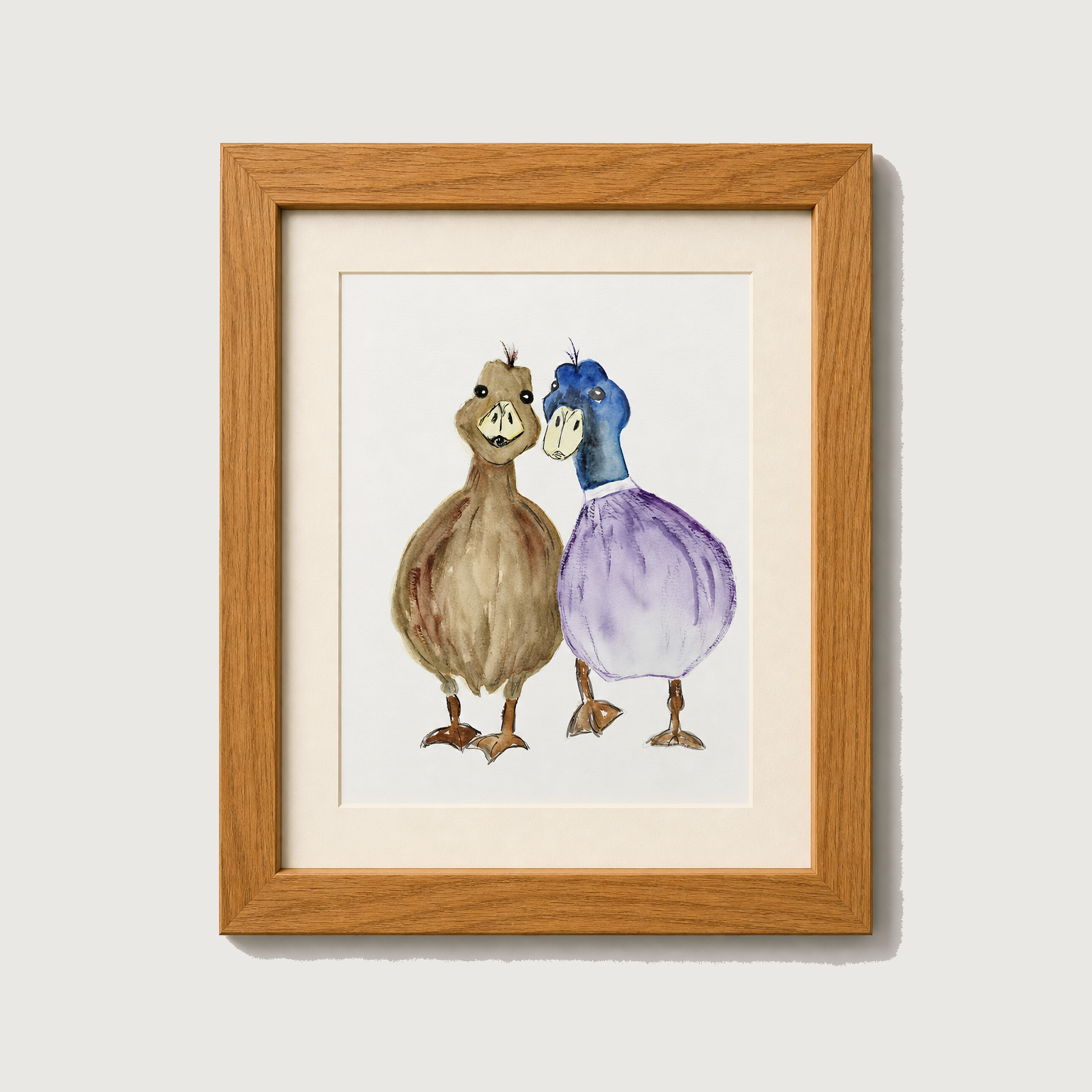 Framed watercolor art print featuring two cartoonish ducks standing side by side, one brown and one blue and purple, set in a light wood frame with a white mat against a light gray wall.