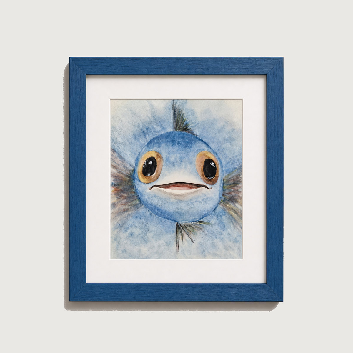 A framed art print featuring a watercolor-style illustration of a blue fish with large eyes, set in a blue frame with a white mat against a light gray wall.