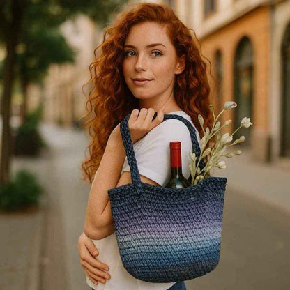 A woman with red hair holds a blue and purple crochet bag, showcasing her vibrant style and unique accessory choice.
