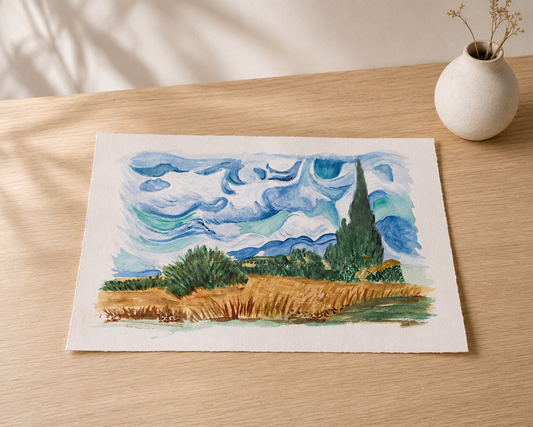 A watercolor landscape art print depicting a blue sky with swirling clouds, green trees, and golden fields, displayed on a light wood table next to a small white ceramic vase.