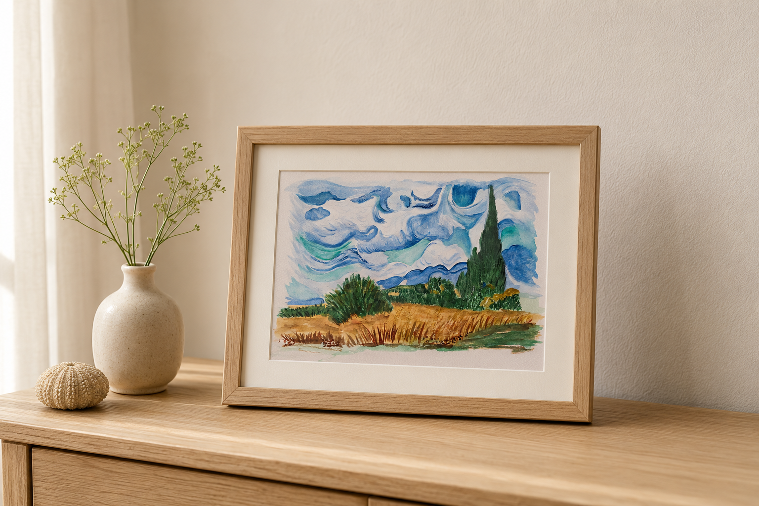 A framed watercolor landscape print featuring a cypress tree and wheat field, styled on a wooden table next to a vase with dried stems in a warm, neutral room.