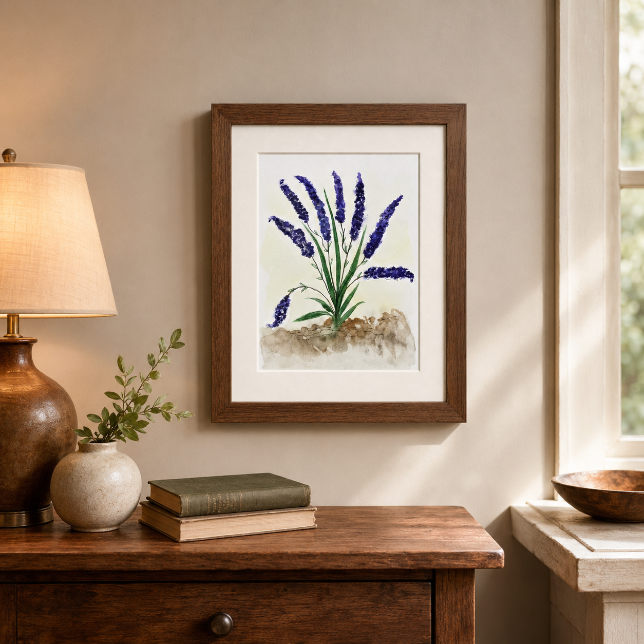 A framed botanical art print of purple lavender flowers is displayed on a beige wall above a wooden console table with a lamp, books, and a small vase in a softly lit room.