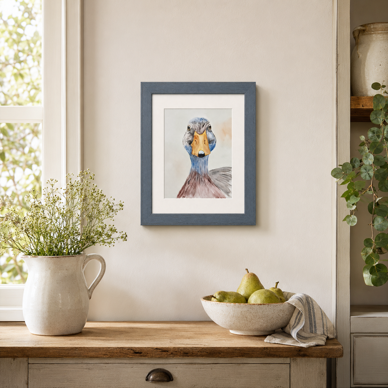 A framed art print of a duck portrait hanging on a beige wall in a styled room setting with a wooden table, a vase of flowers, and a bowl of pears.