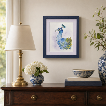 A framed watercolor painting of a blue peacock hanging on a beige wall above a dark wood console table. The room is styled with a brass table lamp, a blue and white ceramic vase with white flowers, a blue and white ceramic bowl, and a large blue and white vase with green branches.
