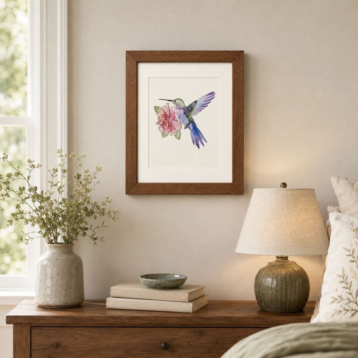 A framed art print of a hummingbird with a pink flower, displayed on a beige wall above a wooden nightstand with a lamp and vase.