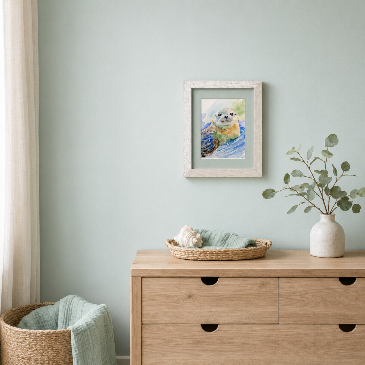 A framed watercolor painting of a seal pupp displayed on a light blue wall above a wooden dresser in a styled room setting.