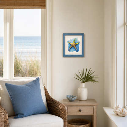 A framed art print of a blue and yellow starfish on a white background, styled in a coastal-themed room above a wooden side table with a vase and beach decor.