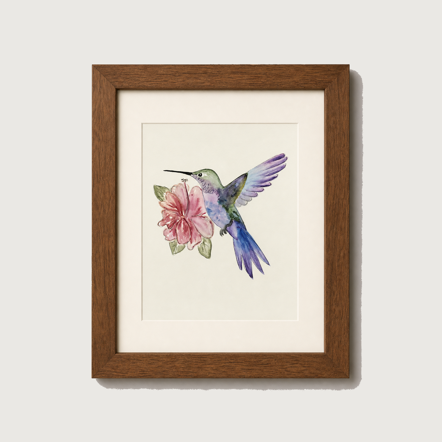 Framed watercolor art print of a hummingbird hovering near a pink flower, set in a brown wooden frame with a white mat against a light gray wall.