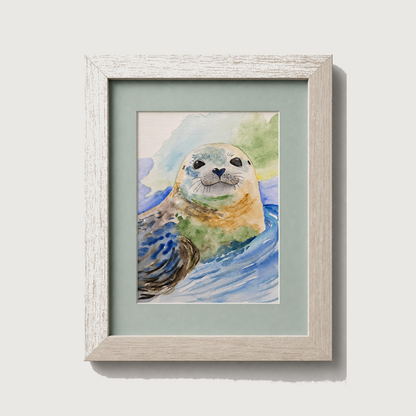 Framed watercolor art print of a seal in blue water, set in a light wood frame with a pale green mat, shown against a neutral light gray background.