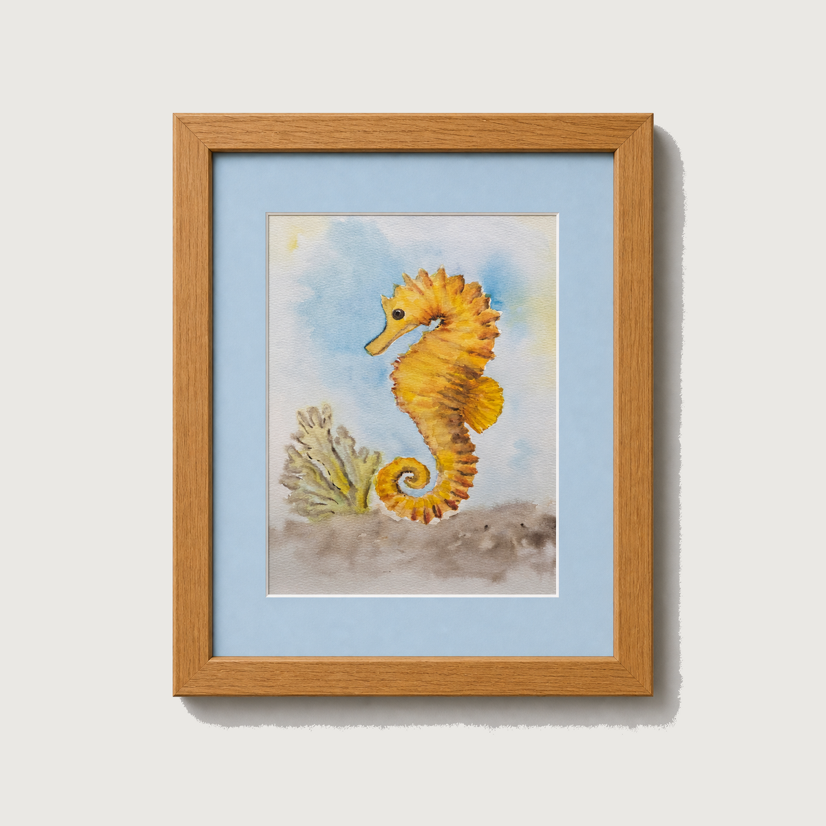Framed watercolor art print featuring a yellow-orange seahorse and coral on a light blue background, set in a light wood frame against a neutral light gray wall.