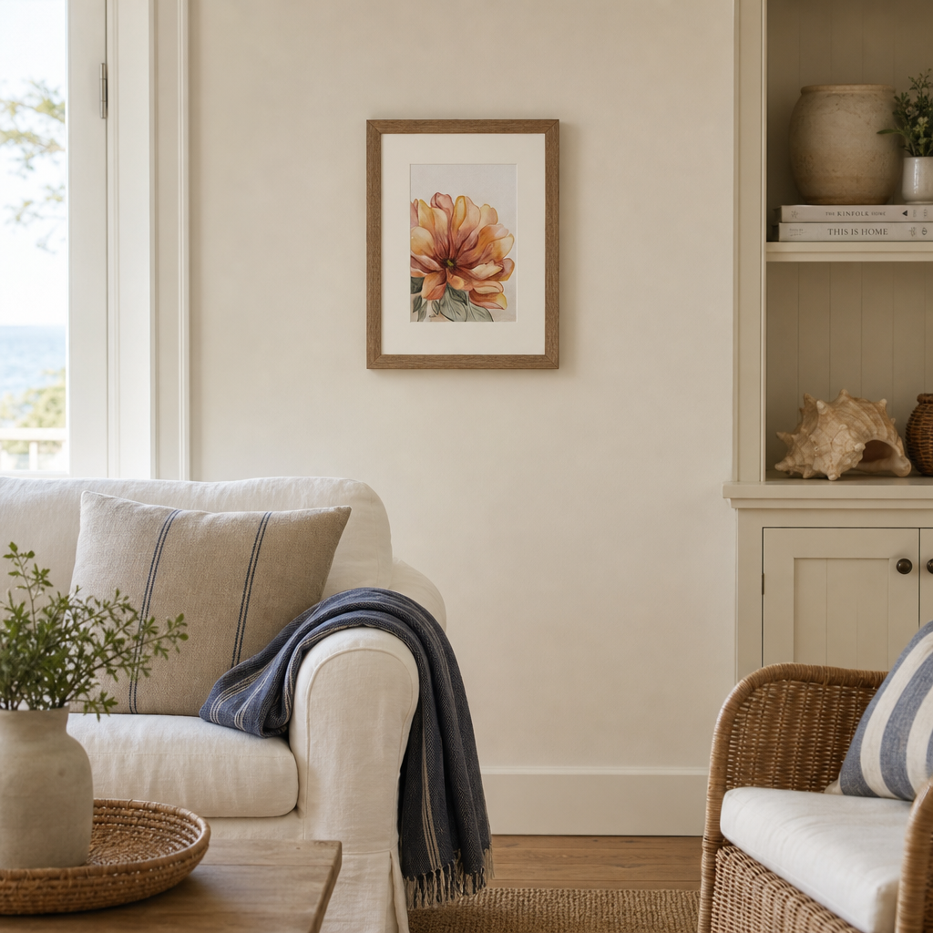 A framed watercolor art print of a warm-toned flower hangs on a cream wall in a styled living room with a white sofa, wicker furniture, and a wooden coffee table.