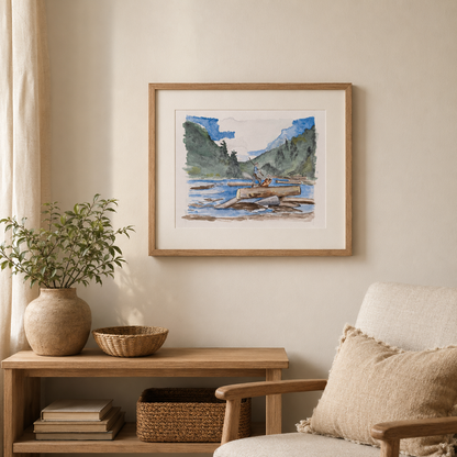 A framed art print depicting a scenic landscape with mountains, water, and a canoe, styled on a beige wall above a wooden console table with a potted plant and a chair.