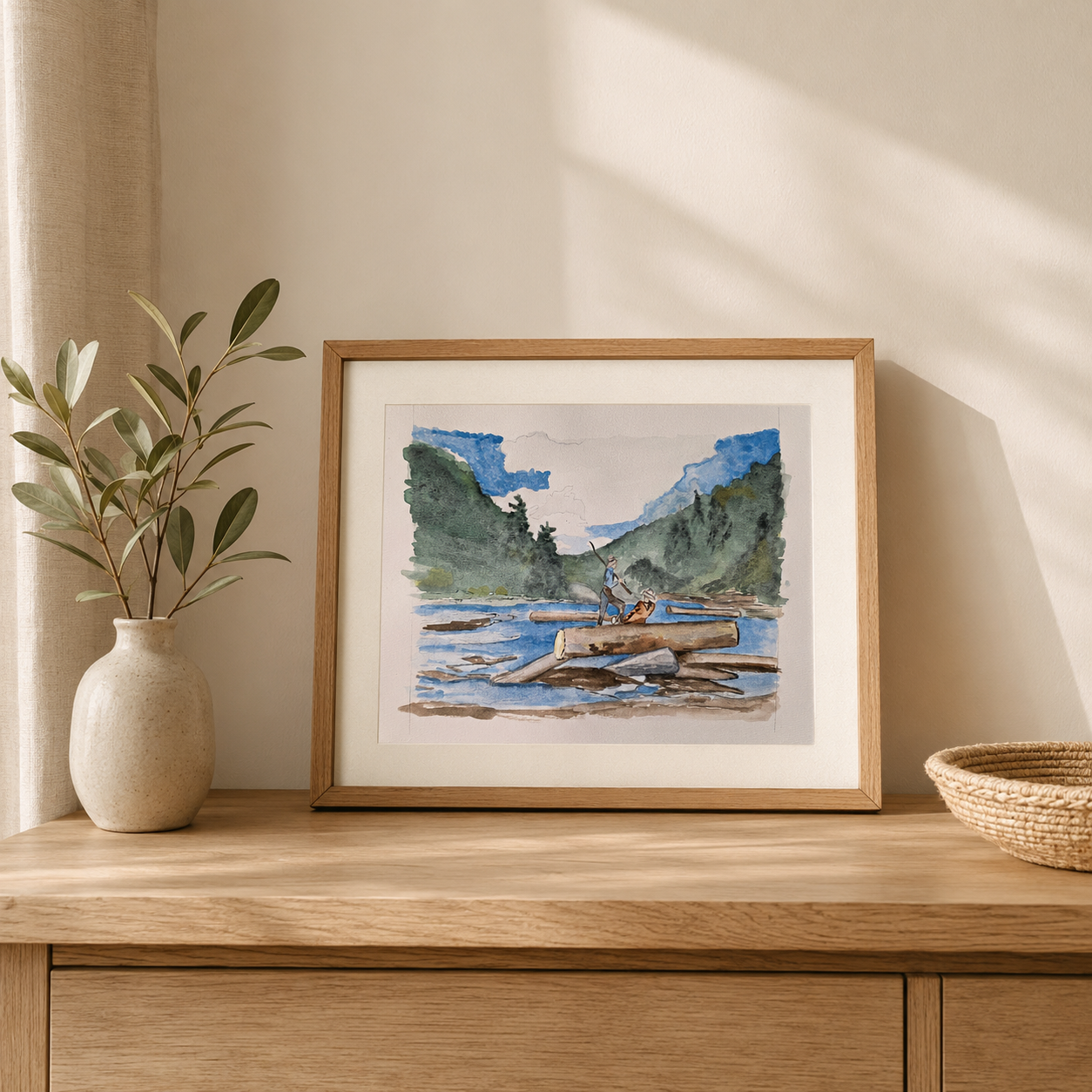 A framed watercolor landscape print of a canoeist on a river, styled on a wooden console table next to a vase with greenery.