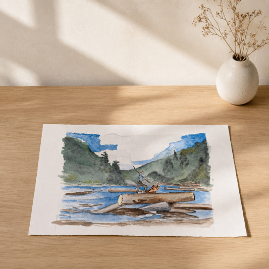 A watercolor art print depicting a person in a canoe on a river with mountains in the background, displayed on a light wooden table next to a small white vase with dried flowers.