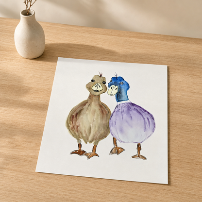 A square art print featuring a watercolor illustration of two ducks standing together, placed on a light wood tabletop next to a small white vase.