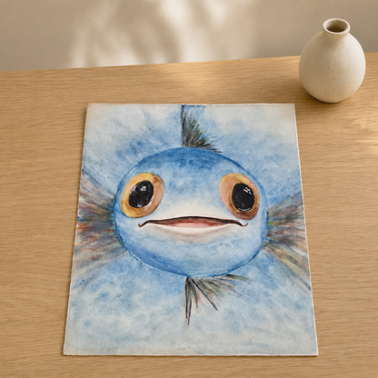 A square canvas print featuring a watercolor painting of a cute blue fish character with large eyes, displayed on a light wooden table next to a small ceramic vase.