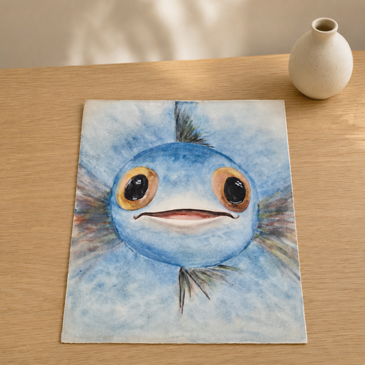 A square canvas print featuring a watercolor painting of a cute blue fish character with large eyes, displayed on a light wooden table next to a small ceramic vase.