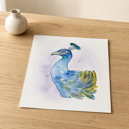 A square watercolor art print of a blue peacock with green and yellow feather details on a white background, displayed on a light wood tabletop next to a small white ceramic vase.