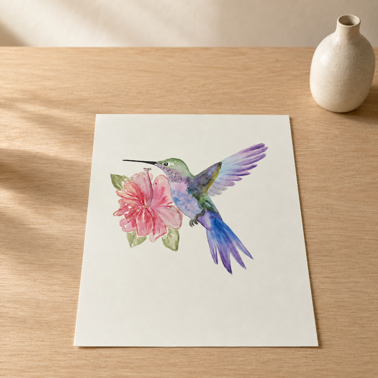 A square art print featuring a watercolor illustration of a hummingbird in shades of purple, blue, and green, hovering beside a pink flower, displayed on a light wood tabletop next to a small ceramic vase.
