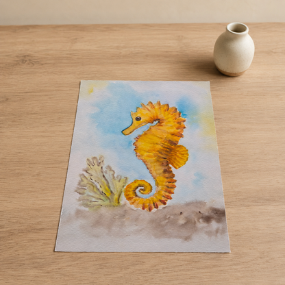 A rectangular art print featuring a painted yellow-orange seahorse with a small coral reef and blue water background, placed on a light wood tabletop next to a small cream vase.