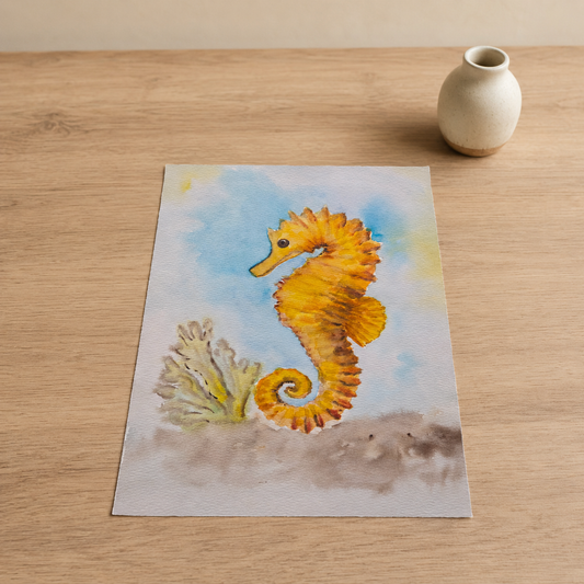 A rectangular art print featuring a painted yellow-orange seahorse with a small coral reef and blue water background, placed on a light wood tabletop next to a small cream vase.