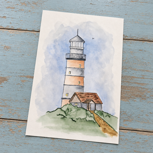 A watercolor art print of a lighthouse on a grassy hill with a small house, displayed on a rustic blue wooden surface.