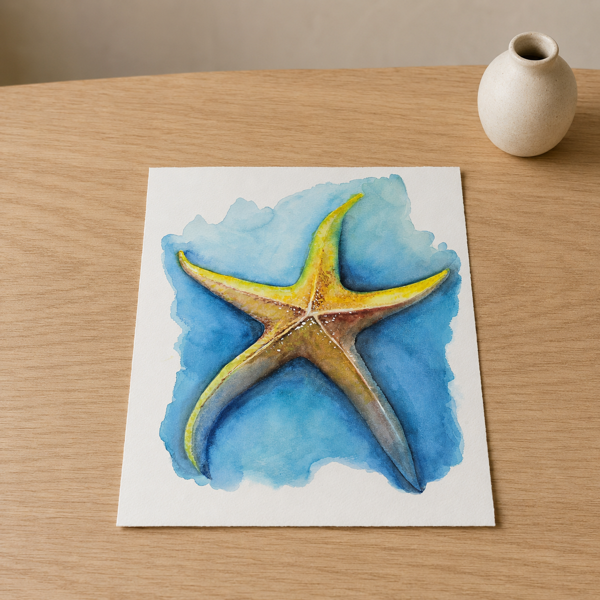 A square art print featuring a painted yellow starfish on a blue watercolor background, displayed on a light wood tabletop next to a small white ceramic vase.